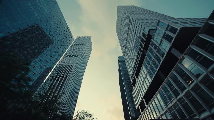 A low-angle tracking shot glides past towering modern glass skyscrapers and trees in a city business district