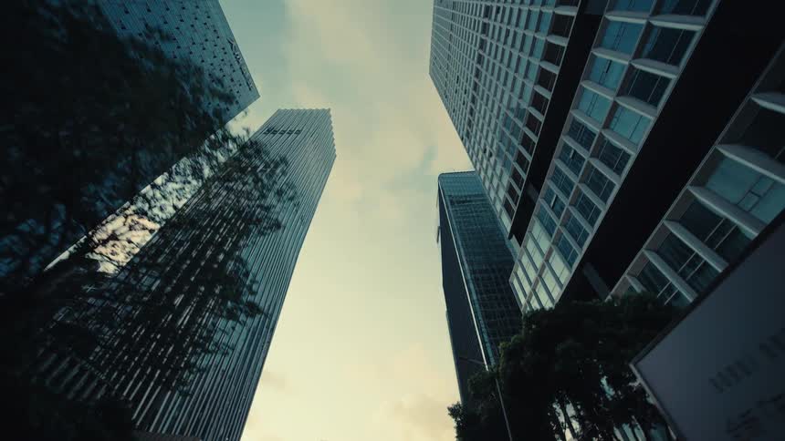 A low-angle tracking shot glides past towering modern glass skyscrapers and trees in a city business district