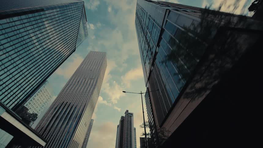 A low-angle tracking shot glides past towering glass skyscrapers in a modern financial district under a cloudy sky