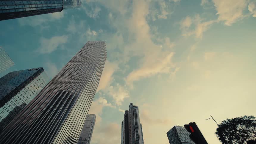 A low-angle tracking shot glides past towering glass skyscrapers in a modern financial district under a cloudy sky