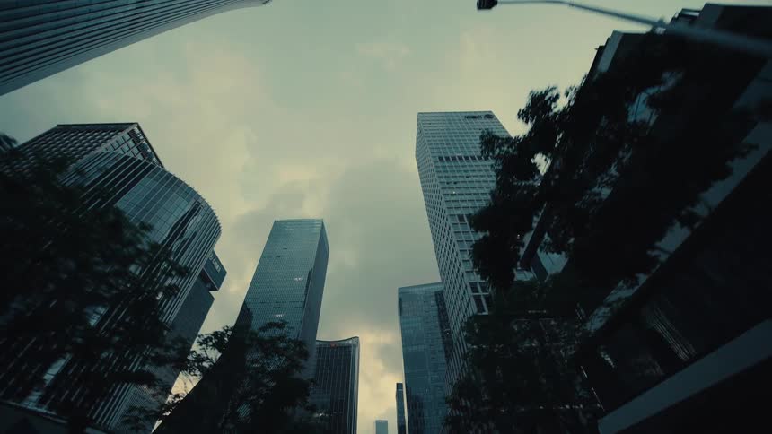 A moody, low-angle tracking shot looking up while driving through a modern financial district with towering glass skyscrapers