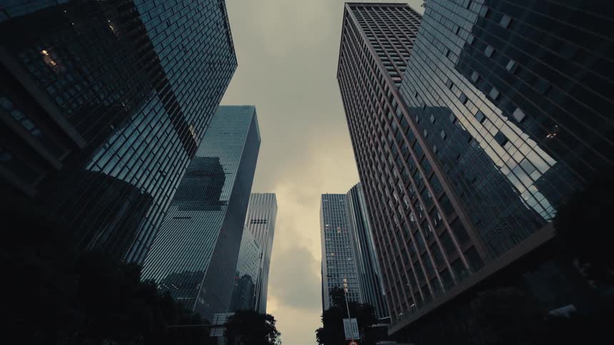 A slow, low-angle tracking shot through a modern city canyon of towering glass skyscrapers reaching for the overcast sky