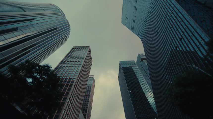 A low-angle driving shot gazes up at modern glass skyscrapers in a bustling city center under an overcast sky