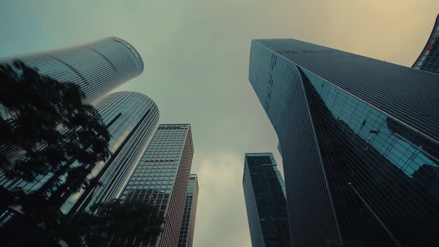 A low-angle driving shot gazes up at modern glass skyscrapers in a bustling city center under an overcast sky
