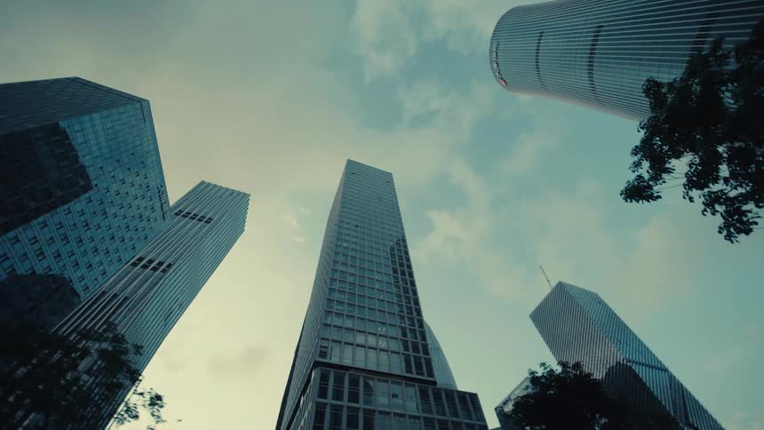 A slow backward dolly shot looking up at an impressive cluster of modern glass skyscrapers reaching into the cloudy sky