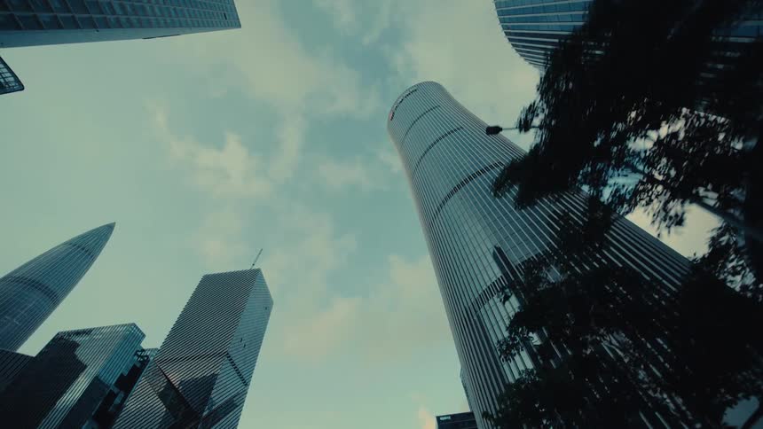 A slow backward dolly shot looking up at an impressive cluster of modern glass skyscrapers reaching into the cloudy sky