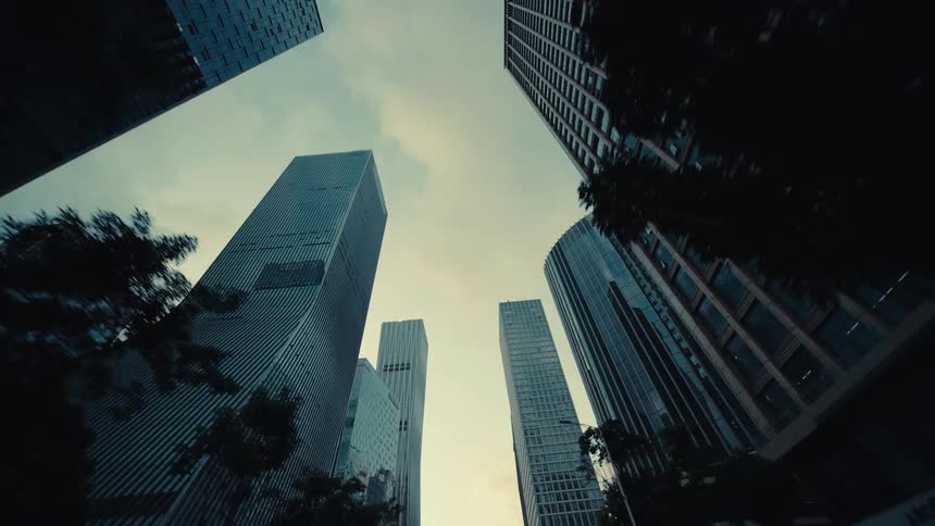 A smooth, low-angle tracking shot moves backward along a city street, looking up at towering modern glass skyscrapers against the sky