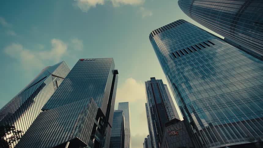 An impressive low-angle tracking shot moves backward through a modern financial district, revealing towering glass skyscrapers against a blue sky