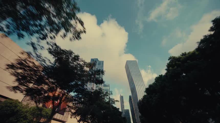 A low-angle reverse dolly shot looking up at modern city skyscrapers and a bright, cloudy sky