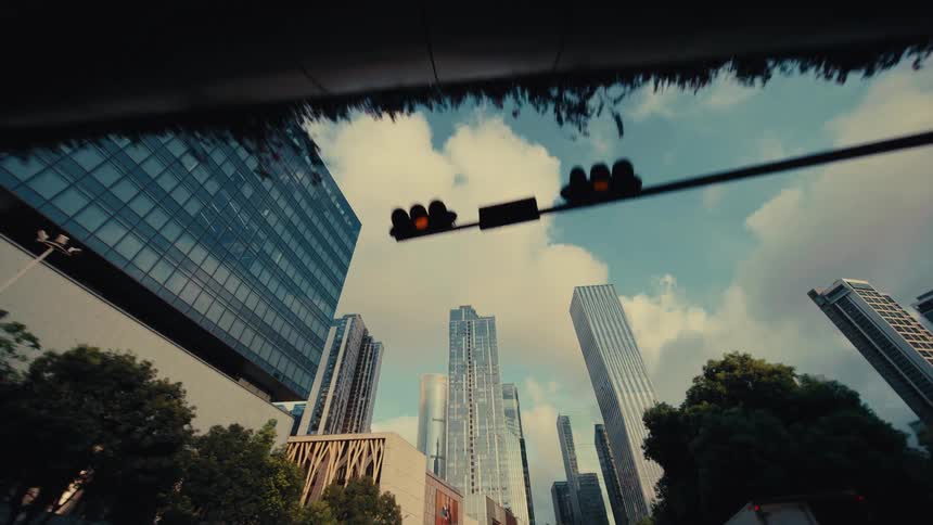 A low-angle reverse dolly shot looking up at modern city skyscrapers and a bright, cloudy sky