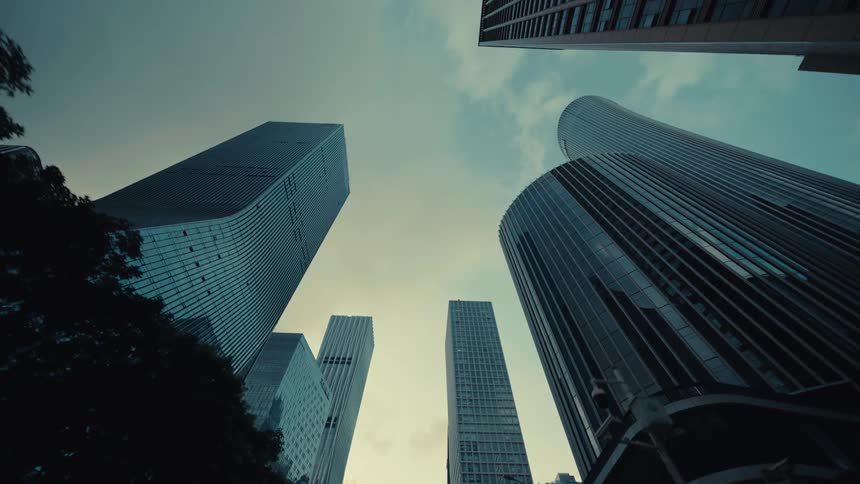 A cinematic low-angle dolly shot moves backward through a modern city, revealing towering glass skyscrapers against an overcast sky