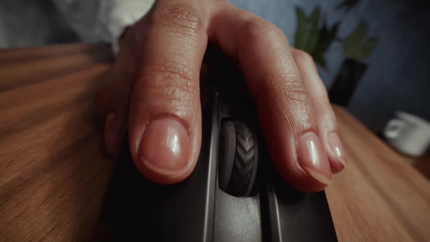 An extreme close-up shot pushes in on a woman's hand as she repeatedly clicks a black computer mouse on a wooden desk