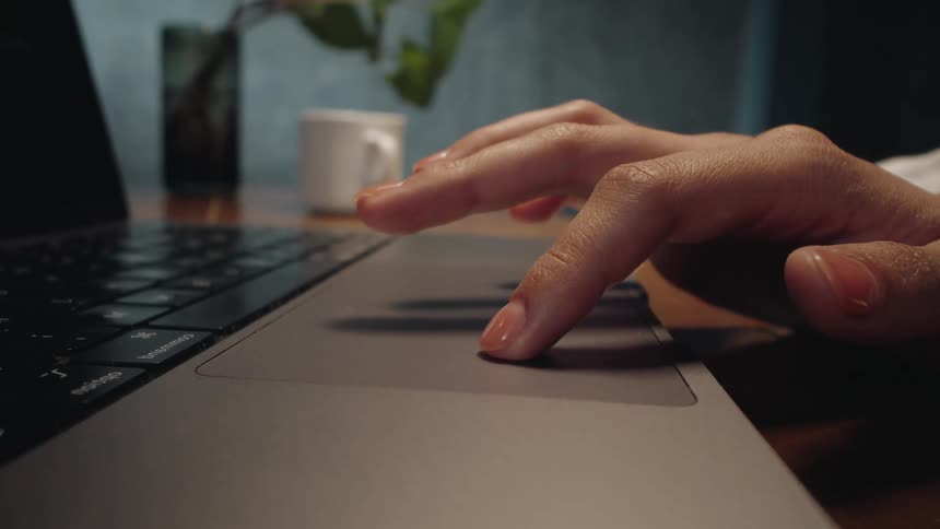 A close-up, slow-motion shot of a hand gently using the trackpad on a laptop at a wooden desk in a dimly lit room