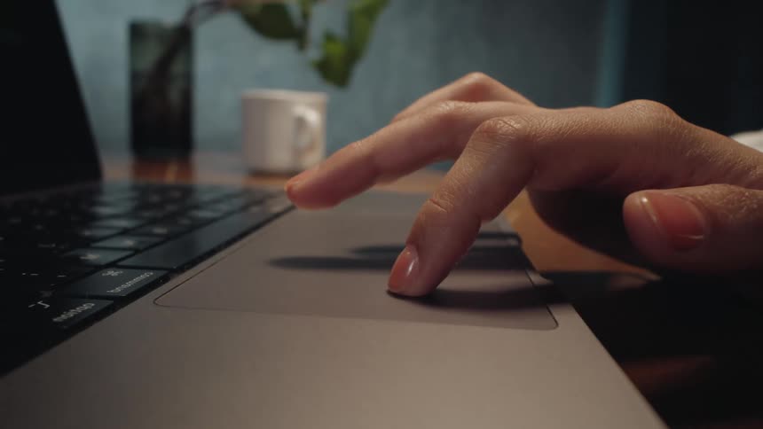 A close-up, dolly-in shot shows a person's fingers using the trackpad on a laptop at a wooden desk