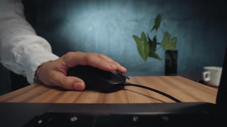 A slow, cinematic push-in shot past a laptop keyboard reveals a person's hand clicking a computer mouse on a wooden desk