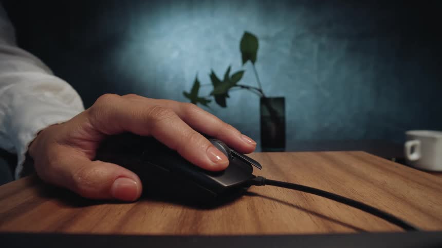 Slow push-in close-up of a hand resting on a computer mouse on a wooden desk next to a laptop in a moody, dark office