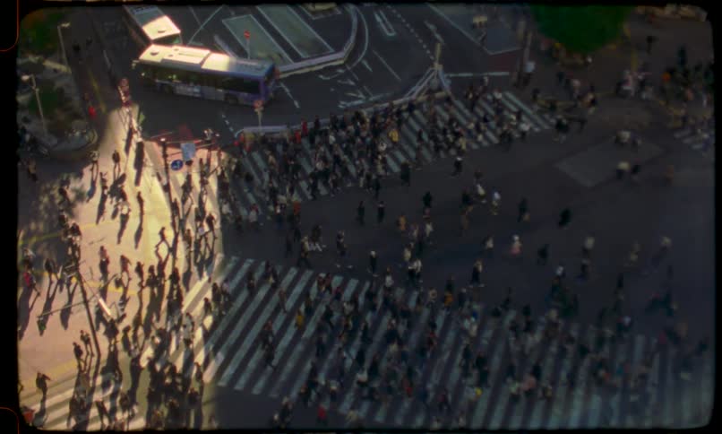 Vintage-style aerial footage captures a massive crowd of pedestrians with long shadows crossing a busy city intersection at golden hour