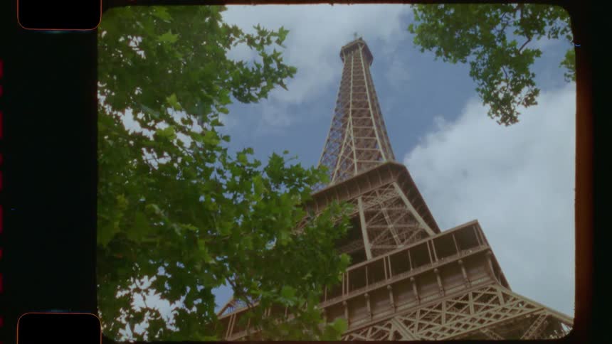 An upward-tilting shot on vintage 16mm film reveals the iconic Eiffel Tower in Paris framed by lush green trees