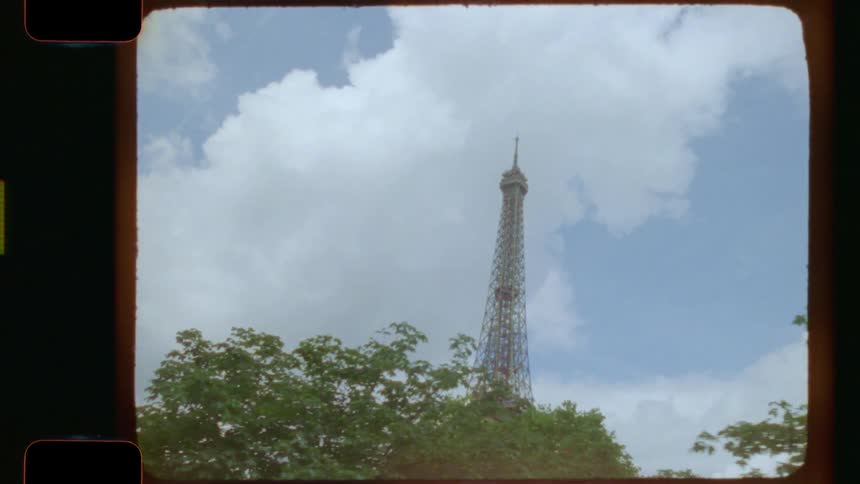 A vintage 16mm film captures a moving shot of the Eiffel Tower emerging from behind lush green trees in Paris