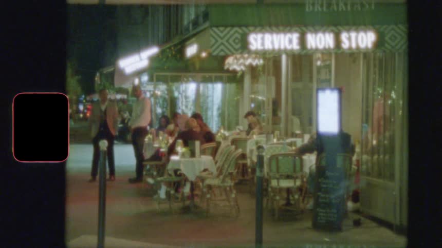 Super 8 film captures the lively atmosphere of a Parisian sidewalk cafe at night as people dine and cars pass by