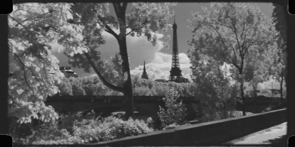A classic, infrared black and white 16mm film captures a picturesque view of the Eiffel Tower in Paris, framed by trees and a bridge