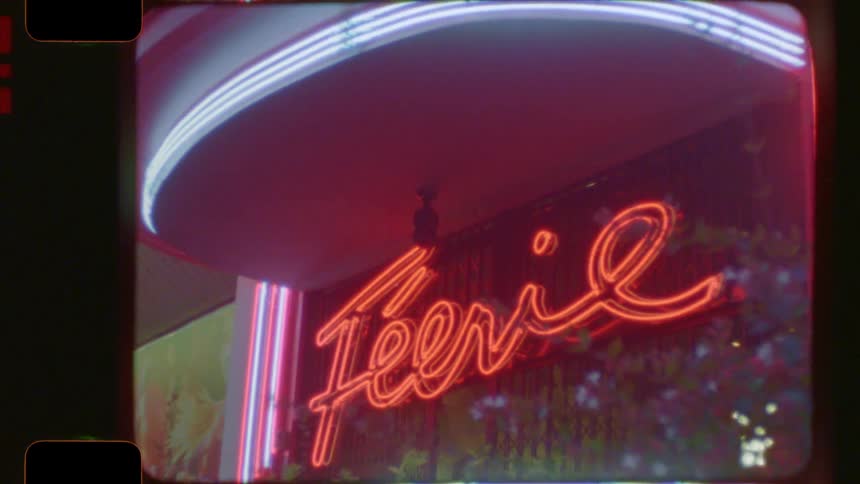 A retro 'Féerie' neon sign blinks and changes colors from green to red on a building at night, captured on vintage 16mm film
