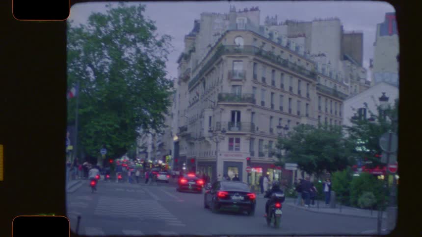 Vintage 16mm film captures an evening street scene in Paris, starting with a brightly lit bistro and pulling back to reveal city traffic