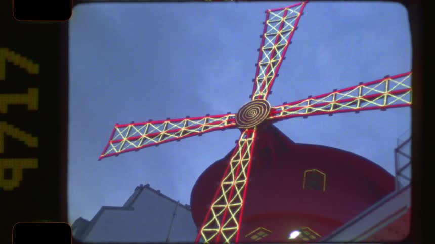 Vintage 16mm film footage captures the iconic red Moulin Rouge windmill illuminated with neon lights at dusk in Paris