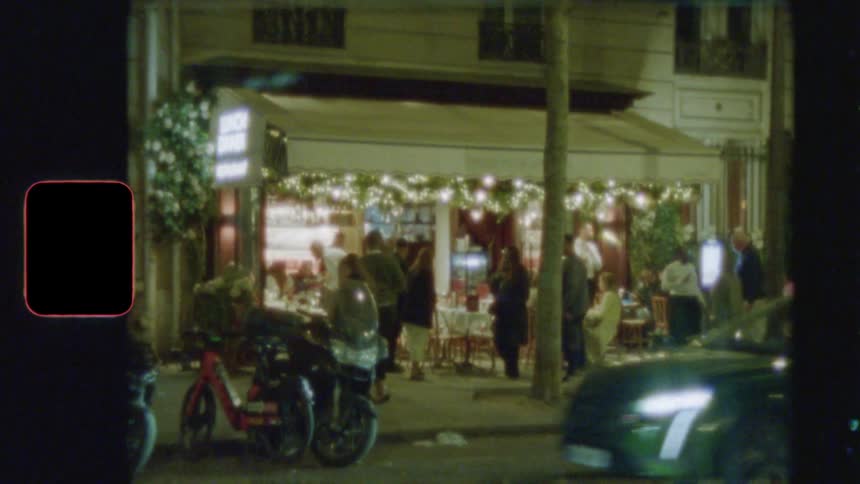 A grainy, vintage-style shot of a beautifully lit Parisian restaurant at night, as people socialize and cars drive by