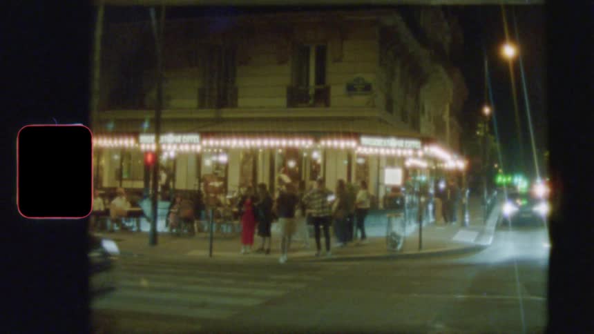 Vintage 8mm film captures a bustling Parisian street corner at night, with people outside a brightly lit cafe as cars drive by