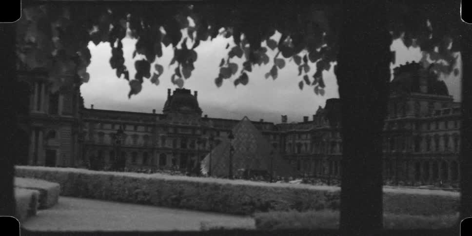 A grainy, black and white 16mm archival view of the Louvre Museum and its iconic pyramid in Paris, France