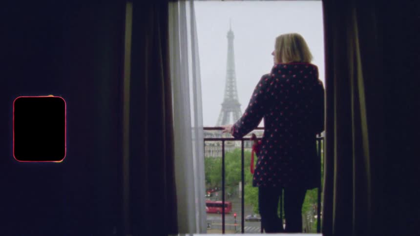 A woman in a stylish coat looks out from a hotel balcony at the Eiffel Tower on an overcast day in Paris, captured on vintage film