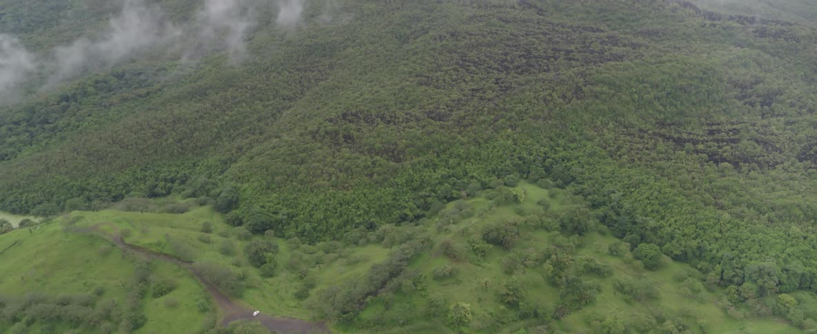 An aerial view tilts up from a white car parked on a dirt road in a lush green valley to reveal the vast, misty, tree-covered mountainside