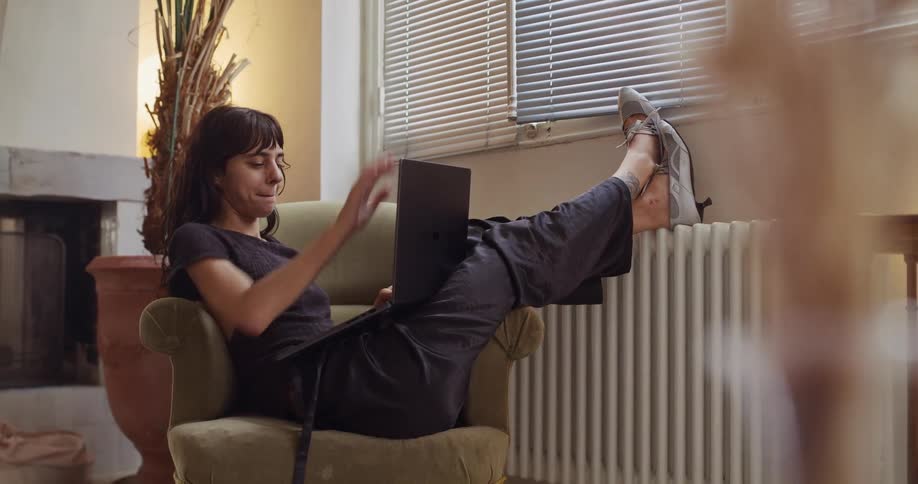 A relaxed woman finishes working on her laptop while sitting in a cozy armchair, closing it with a smile to unwind