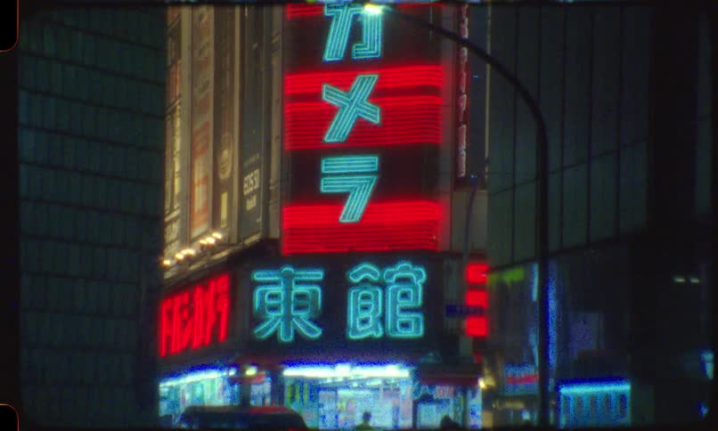 A grainy, vintage film shot pans up a building revealing vibrant Japanese neon signs on a Tokyo street at night