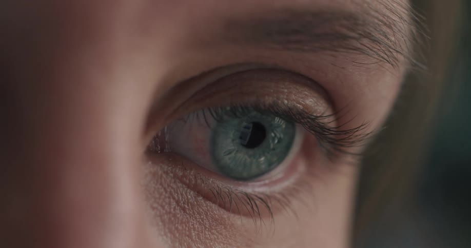 An extreme close-up of a blue-green eye blinking and looking around, with a screen reflected in the pupil