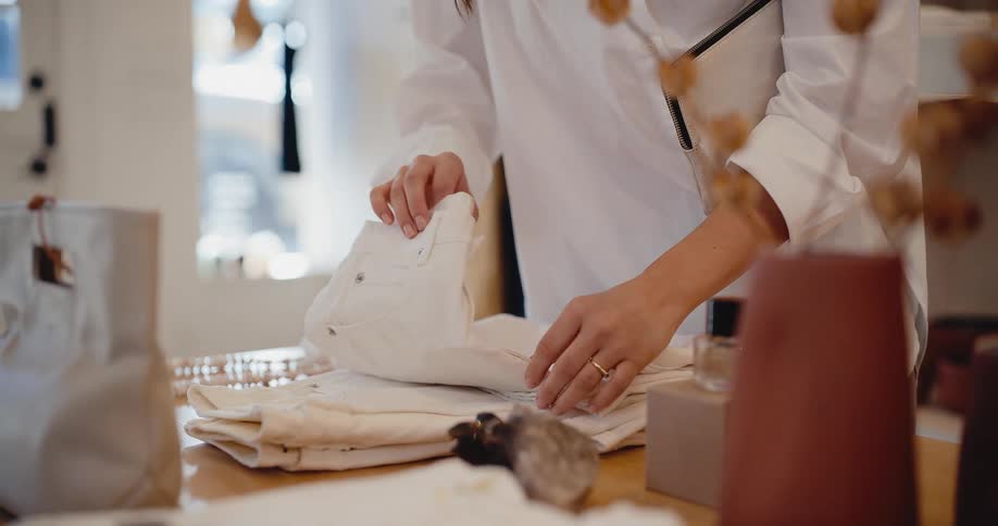 A woman in a white shirt carefully folds a pair of white jeans on a wooden table in a chic, sunlit boutique