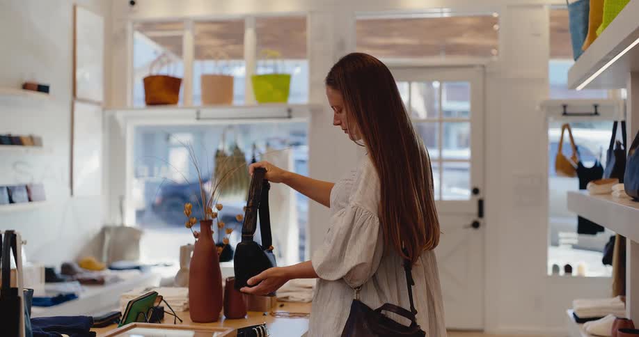 A young stylish woman inspects a black designer handbag while shopping in a modern sunlit boutique