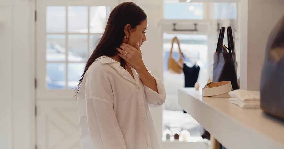 A stylish young woman in a white shirt thoughtfully browses accessories in a bright, modern clothing boutique