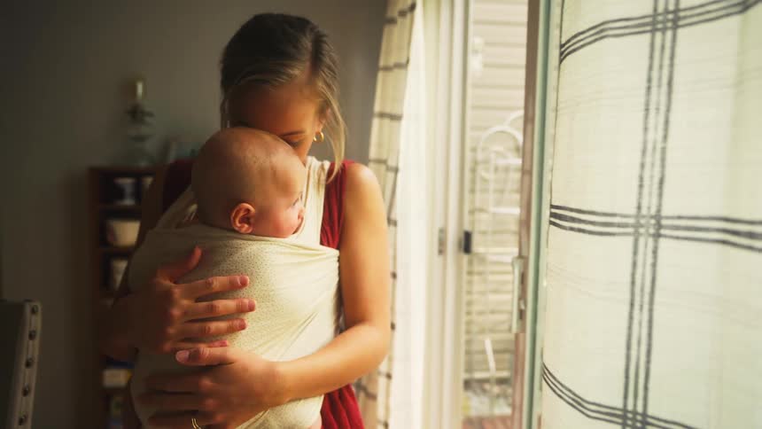 A young mother holding her baby in a sling looks out the window before giving the infant a loving kiss on the head