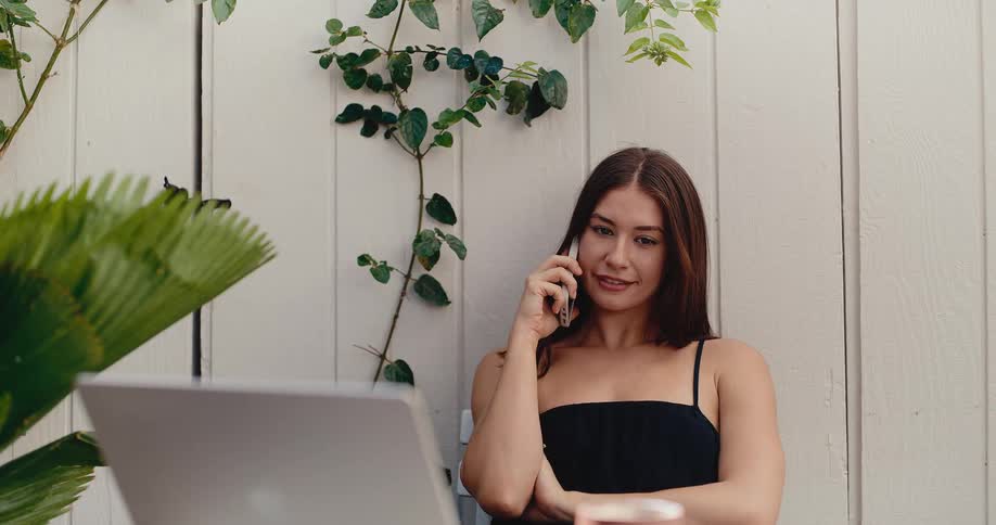 A young woman talks on her smartphone while working remotely on her laptop from a beautiful outdoor patio