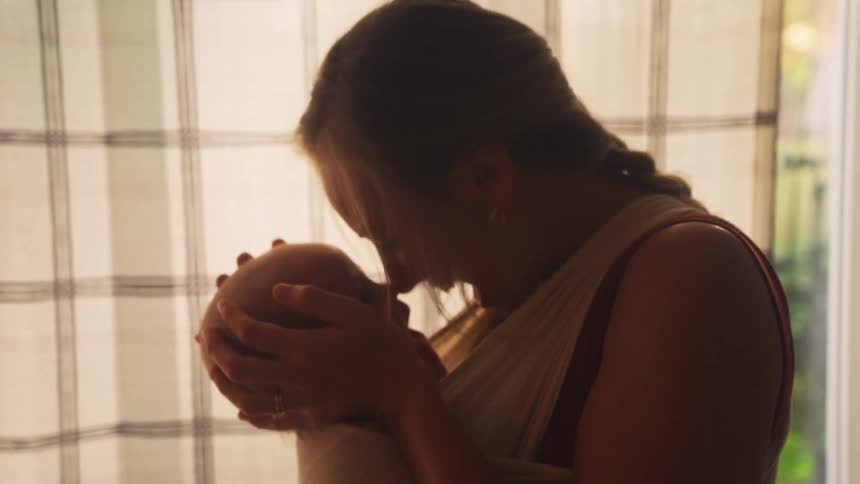 A silhouetted mother smiles and gazes lovingly at her newborn baby held close in a carrier