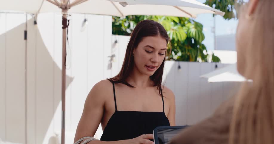 A smiling young woman in a black dress talks to a friend on a sunny patio before opening her wallet