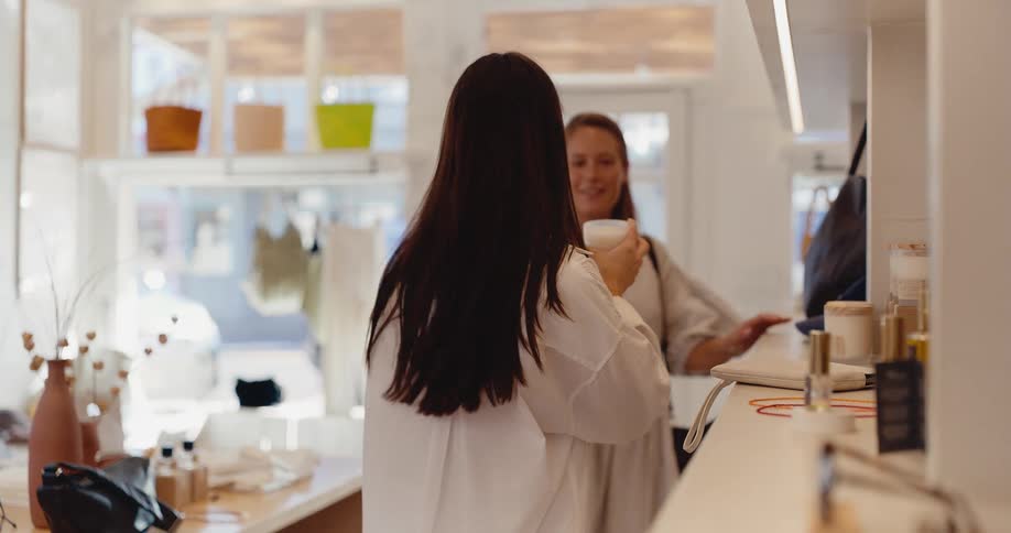 A young woman smells a scented candle while shopping for beauty products in a bright and airy boutique as the friendly owner assists her