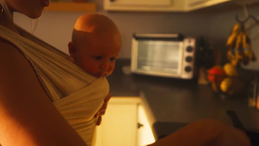 A multitasking mother cooks ground beef in a skillet on the stovetop while holding her baby in a carrier