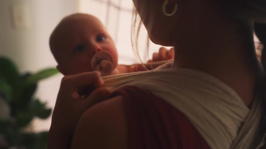 A mother tenderly holds her newborn baby with a pacifier, who is nestled securely in a baby sling in a dimly lit room
