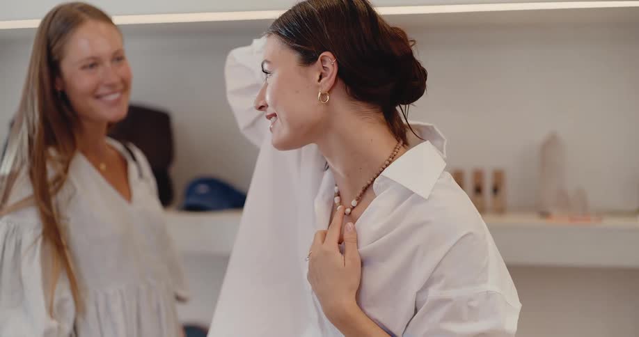 A smiling young woman tries on a beaded necklace while shopping with her friend in a stylish boutique