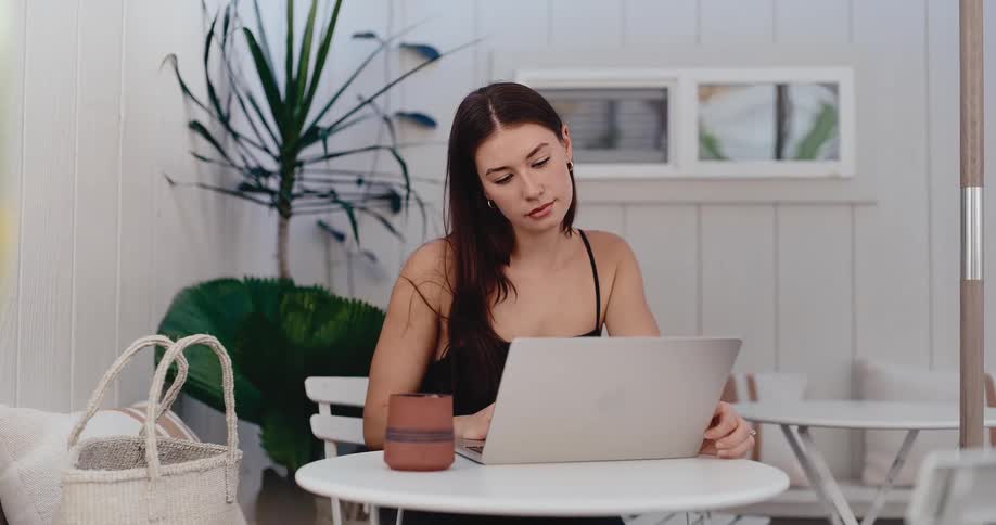 A young woman with dark hair works on her laptop at a bright, minimalist cafe, smiling as she looks at the screen