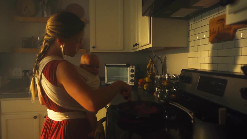 A young mother multitasks by cooking dinner on the stove while holding her infant in a cozy baby wrap