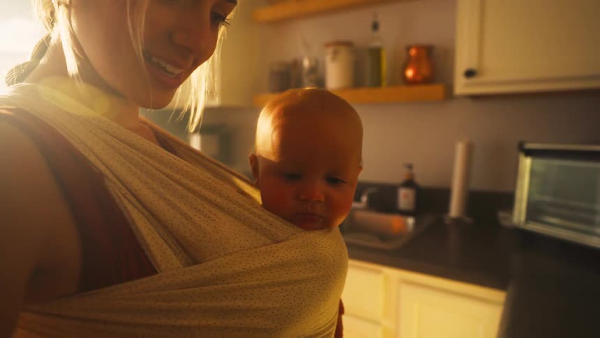 A loving mother holds her newborn baby close in a sling while standing in a sunlit kitchen